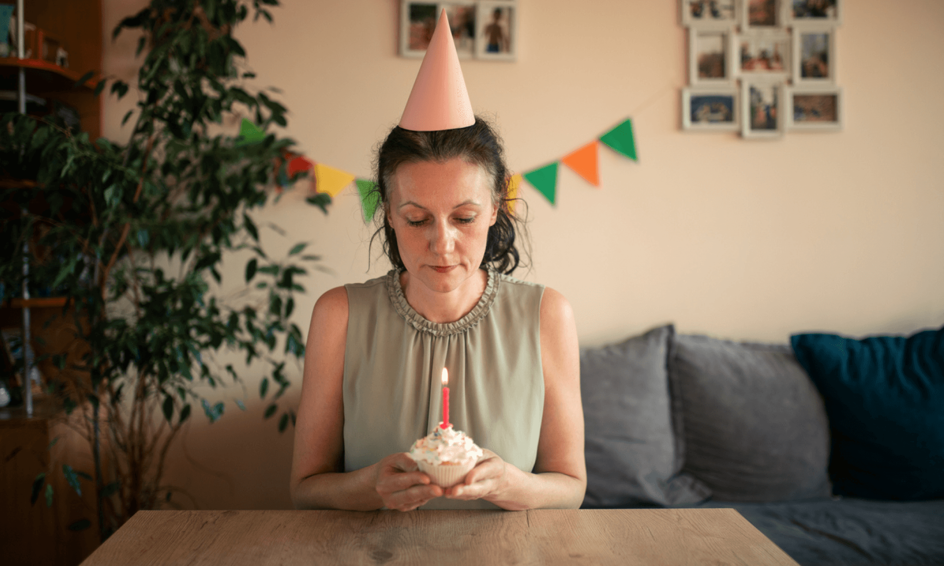 A woman sitting at a table wearing a birthday hat and holding a cupcake with a birthday candle in it